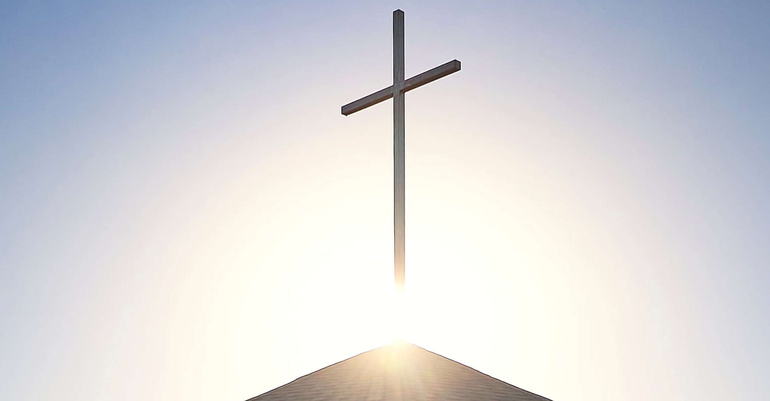 A large cross atop a rooftop is silhouetted against a bright, glowing sunrise, conveying a sense of peace and spirituality under a clear blue sky.