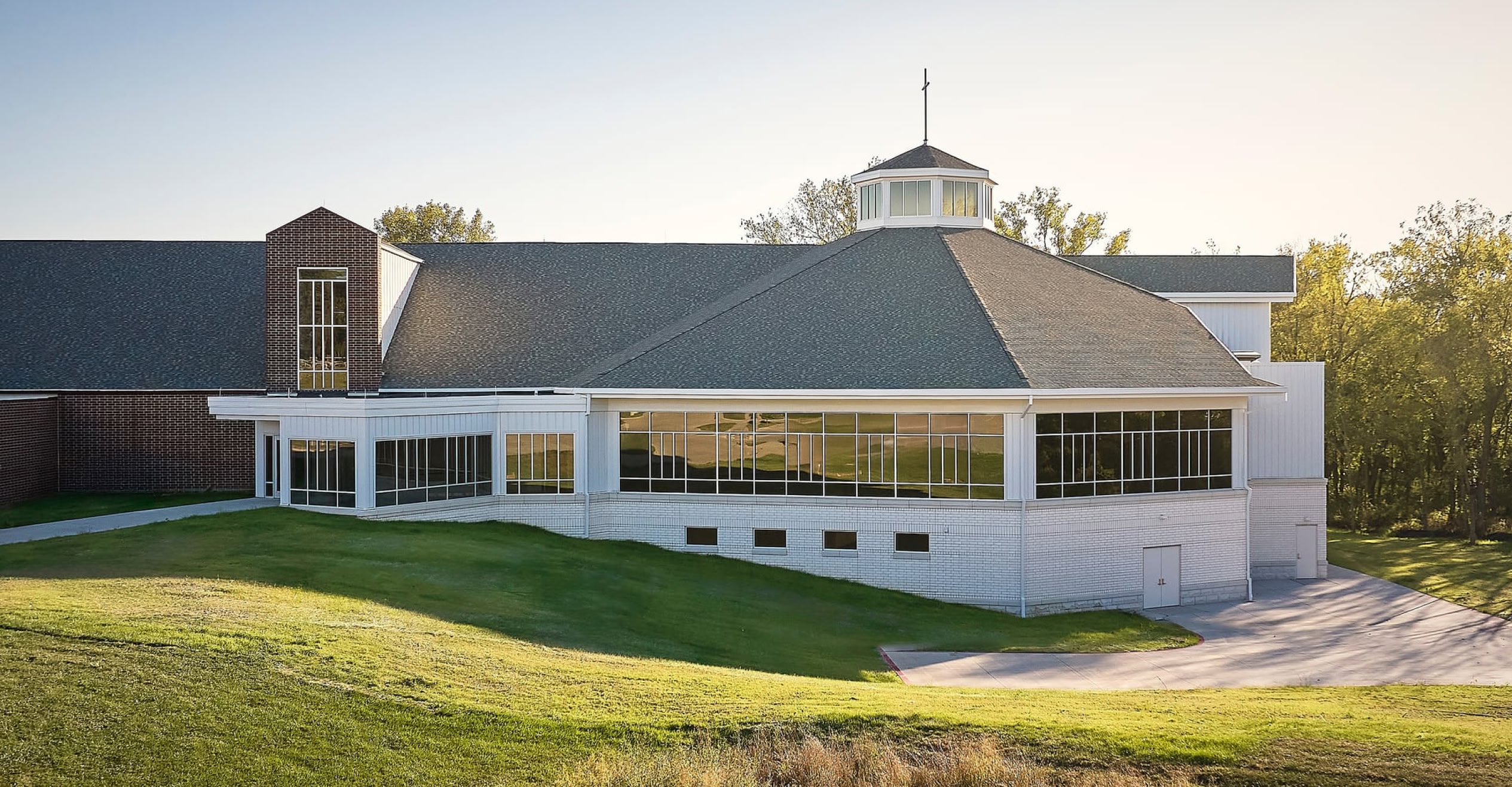 A modern church building with large windows and a central steeple surrounded by greenery.
