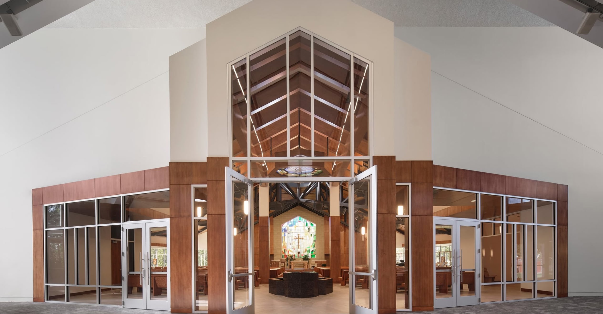 Open church entrance with large glass doors and wooden frame, leading to an interior with high vaulted ceiling, stained glass window, and rows of seating.