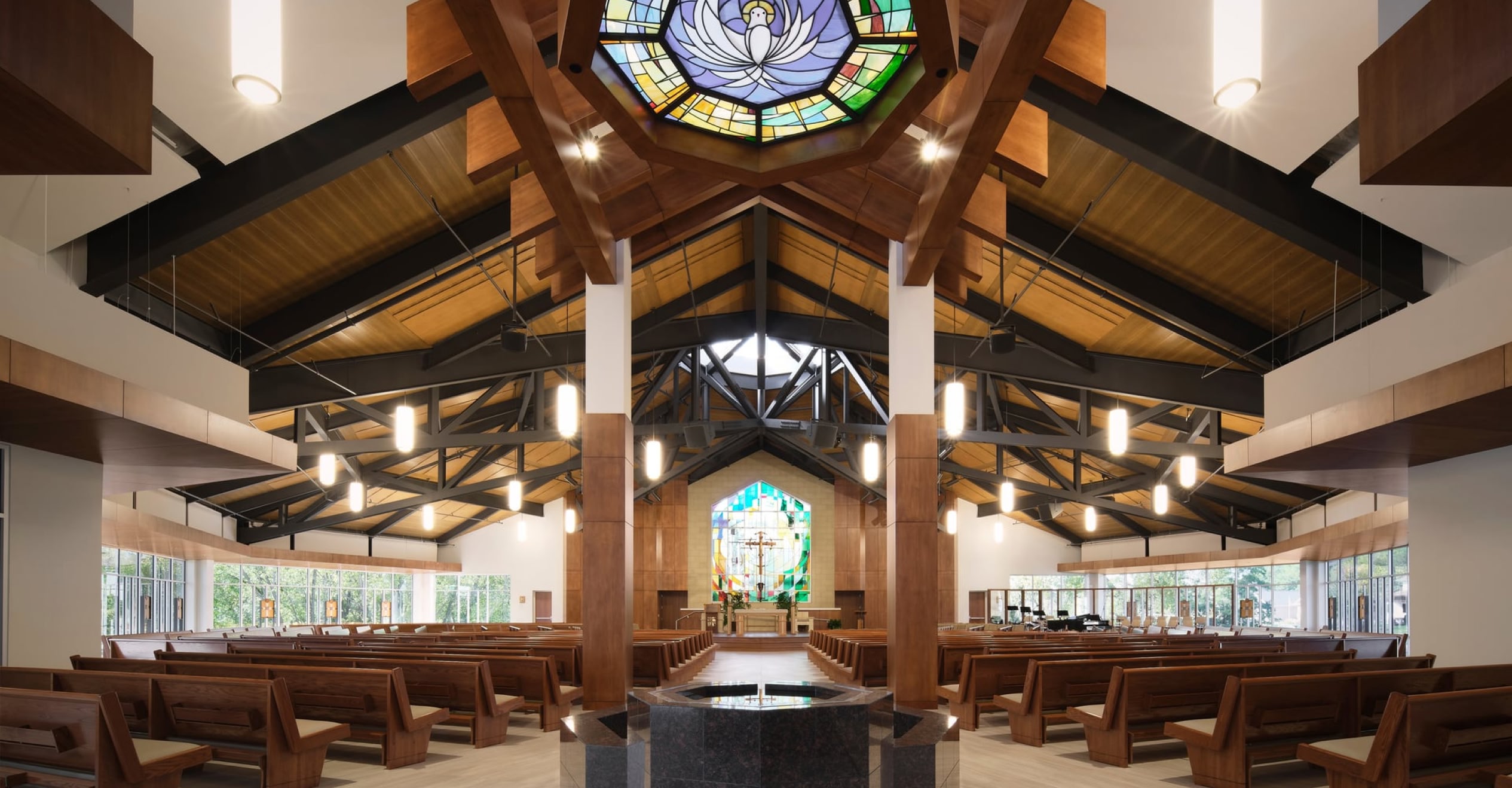 Spacious modern church interior featuring wooden pews, vaulted ceiling with a colorful stained glass dome, and an altar with a large stained glass window.