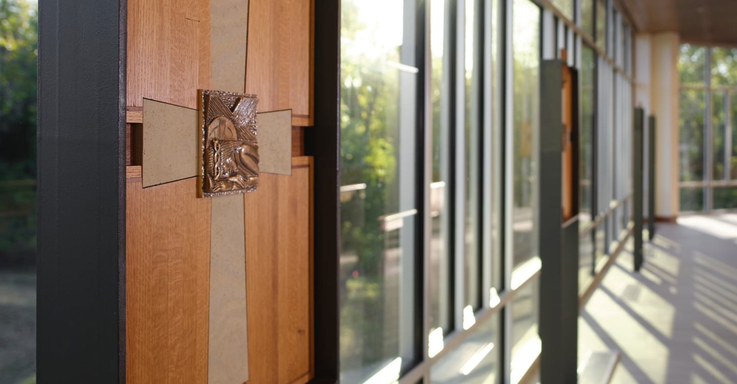 A sunlit hallway with large glass windows casts shadows on the floor. A wooden panel with decorative detail is prominently displayed on the left.