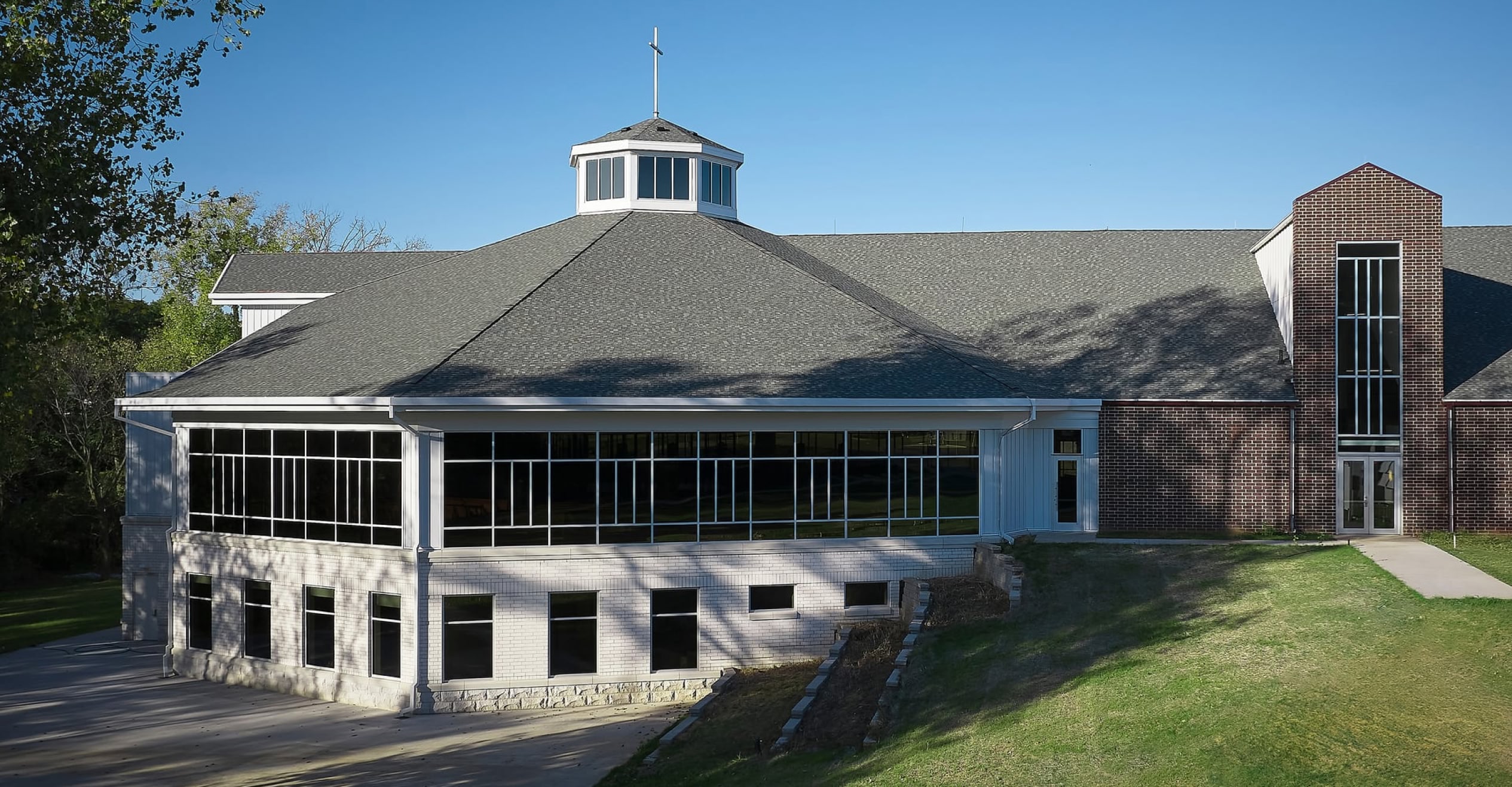 A modern church with large windows and a cross-topped cupola sits on a sunny day. Brick and white exterior, surrounded by green grass and trees.