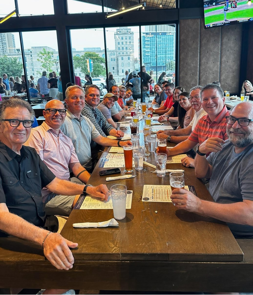 A group of smiling people seated at a long restaurant table with drinks and menus, enjoying a casual gathering. Large windows provide a cityscape view.