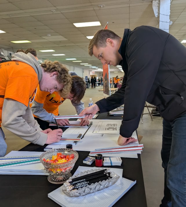 Three people focus on architectural plans at a table. Two wear orange shirts and use rulers, while one instructs.