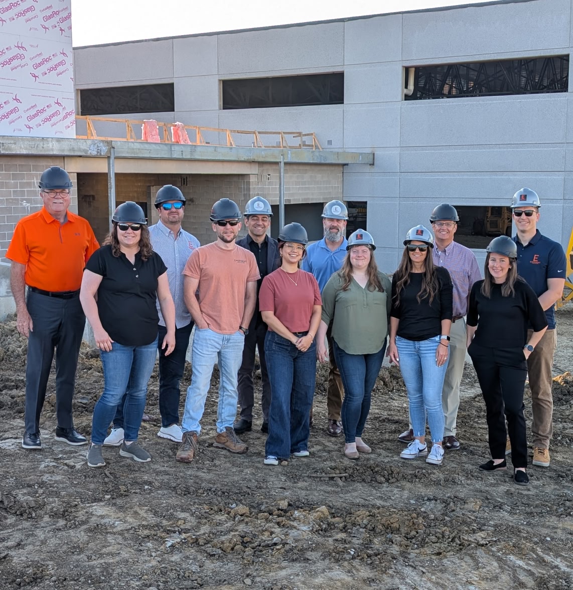 A group of eleven people in hard hats stand on a construction site in front of a partially built structure, smiling