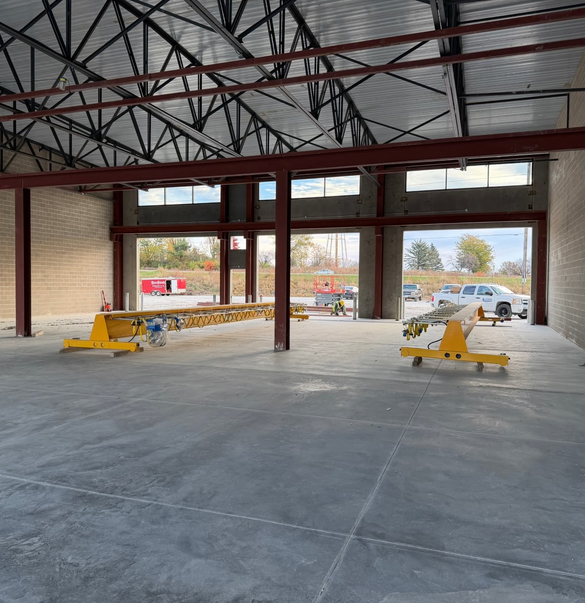 Spacious construction site interior with bare concrete floor and steel beams. Large windows reveal trucks and autumn trees outside