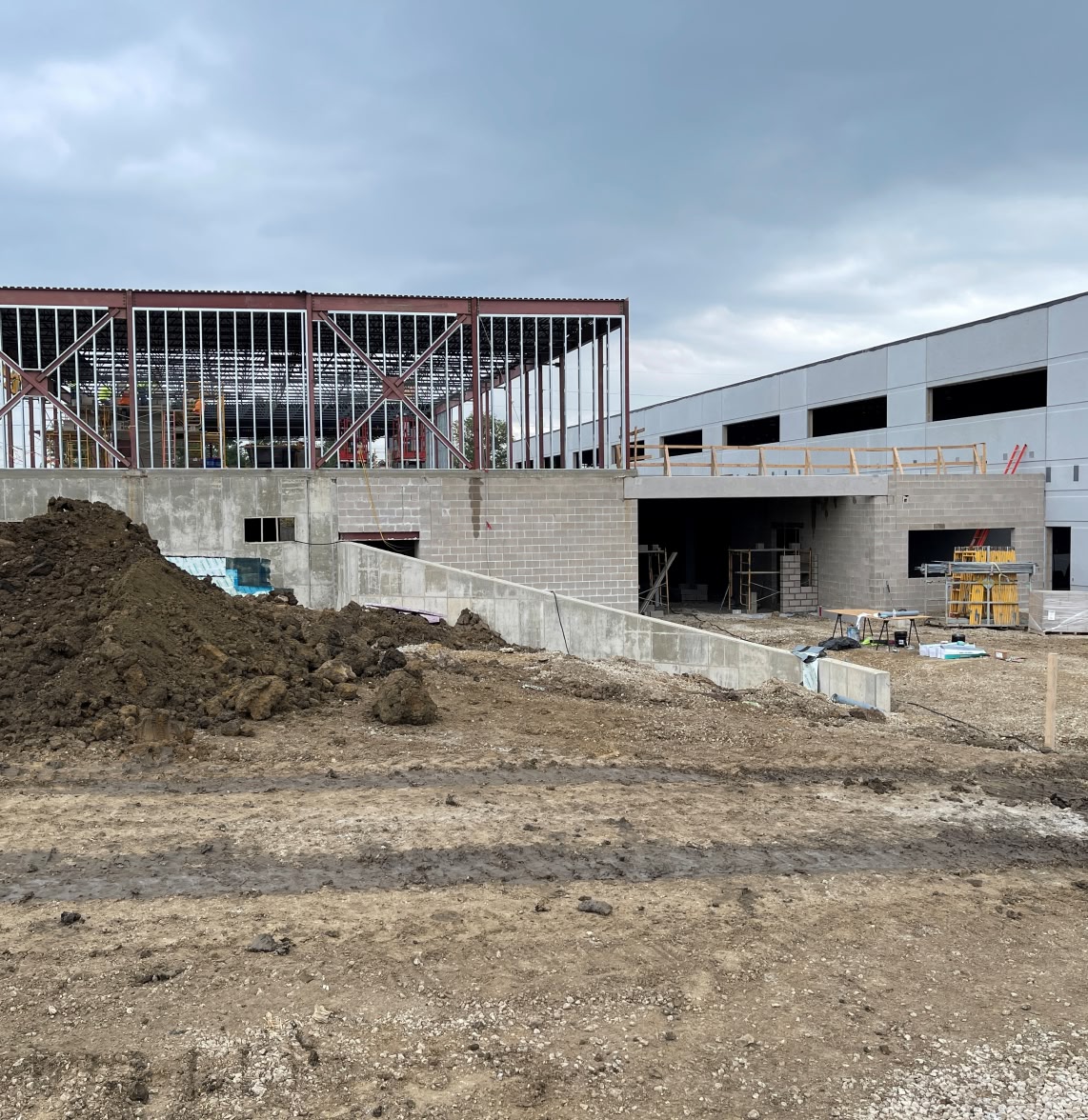 Construction site with a partially built structure featuring steel framing and concrete walls. Piles of dirt and building materials are scattered under a cloudy sky.