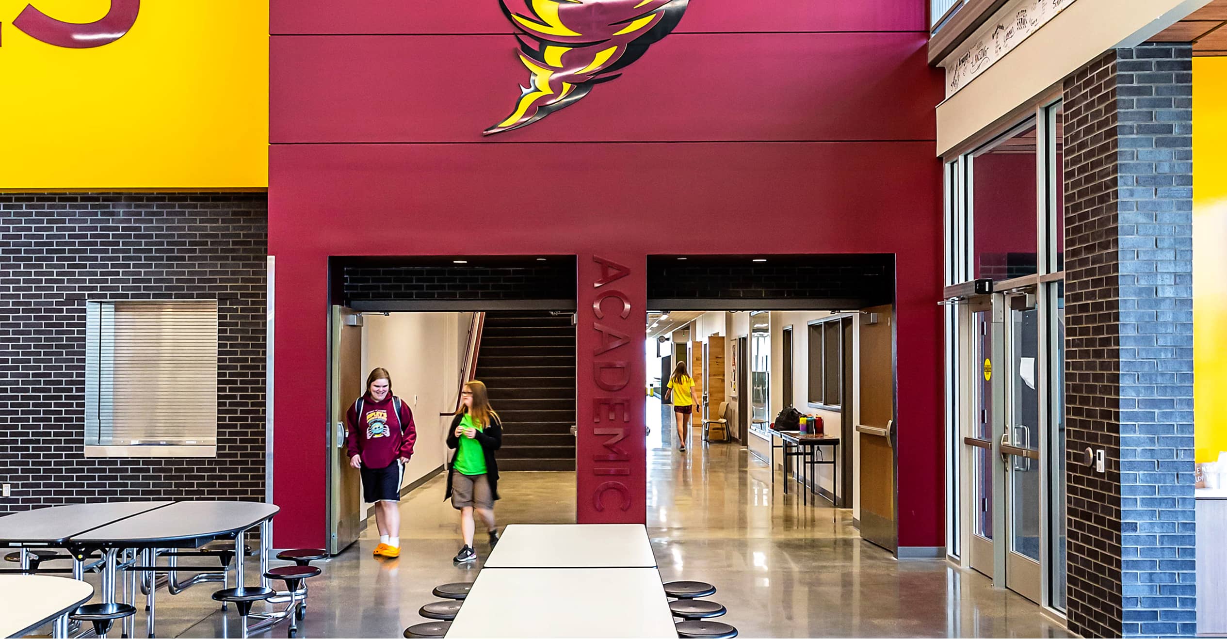 Two students walk through a school hallway labeled "Academic," featuring maroon and yellow walls and a ceiling logo.