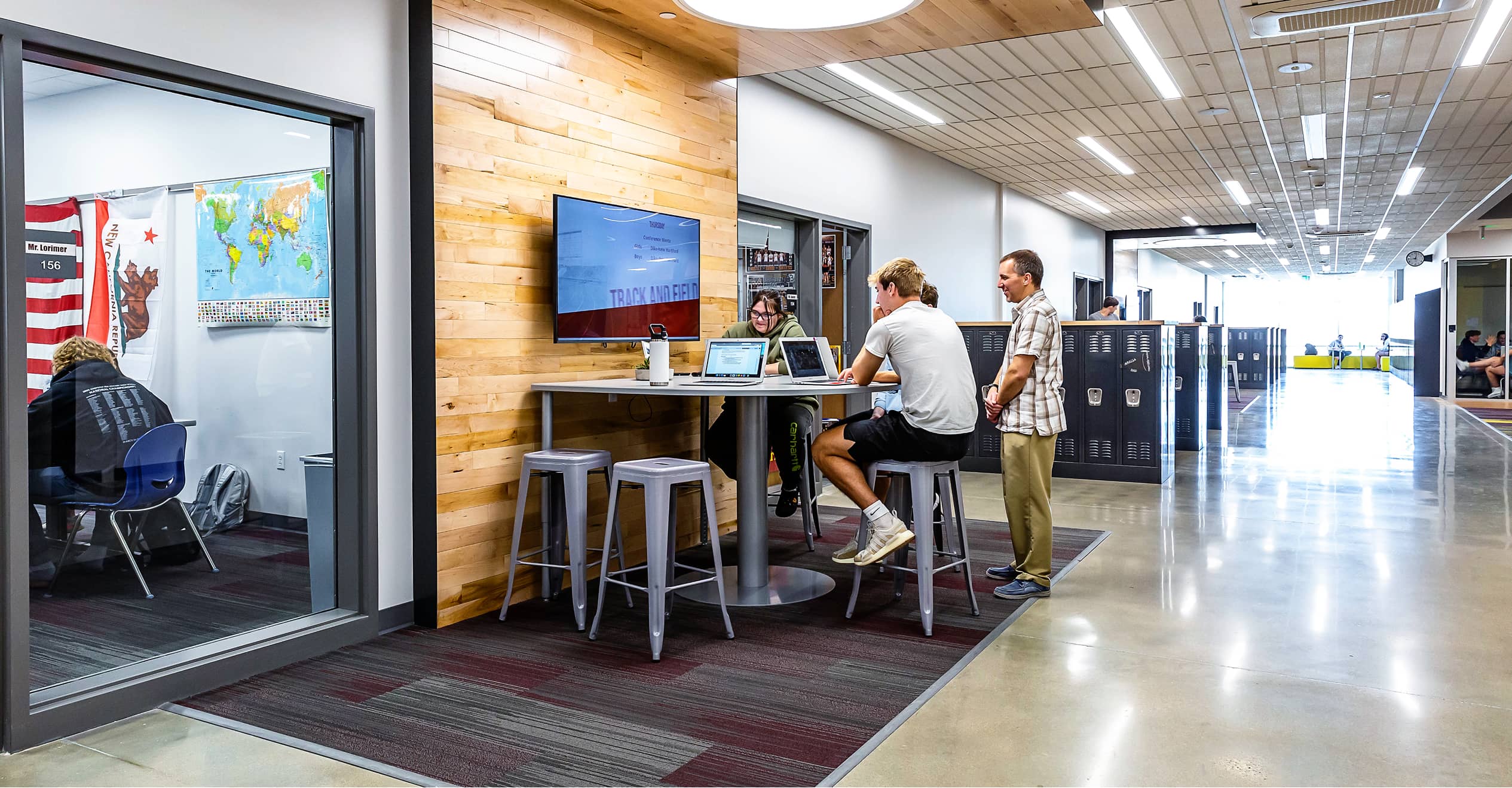 Modern school hallway with students and a teacher at a high table using laptops. Inside a glass-walled room, flags and maps are visible.