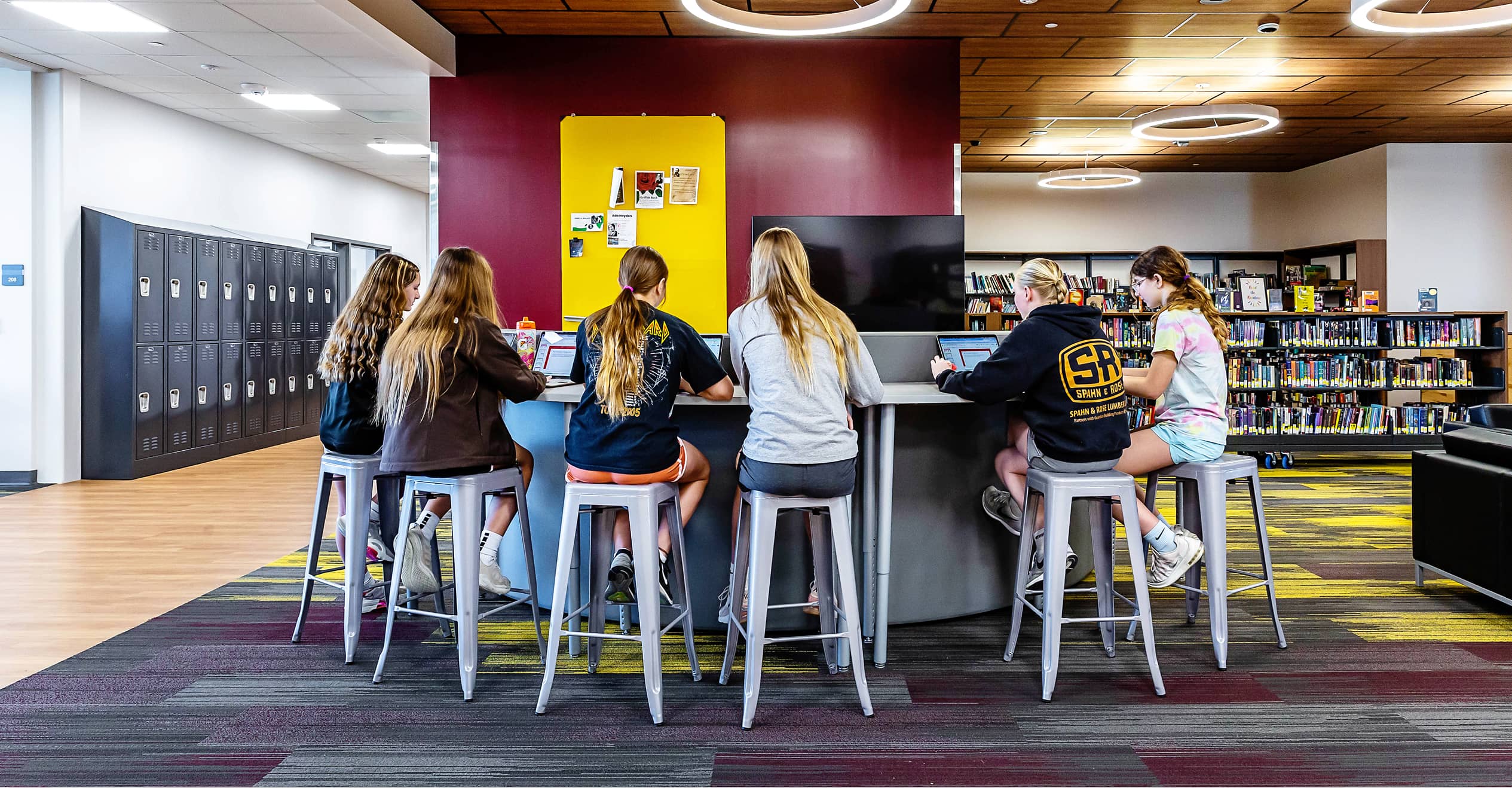 A group of six students with long hair sit on stools around a high table in a modern library. Behind them are bookshelves and lockers