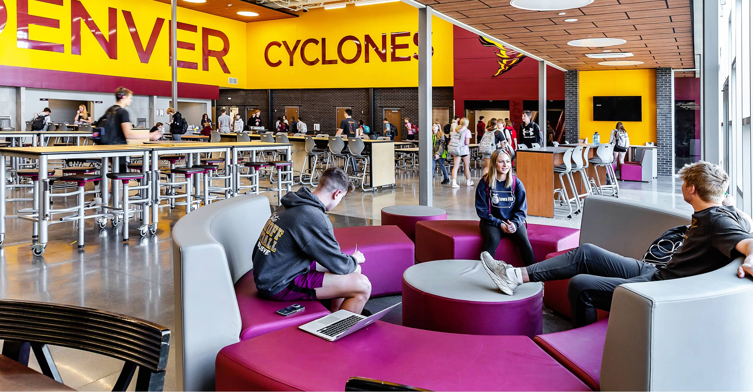 A modern school cafeteria with large "Denver Cyclones" signs. Students in casual wear sit on colorful, circular seating