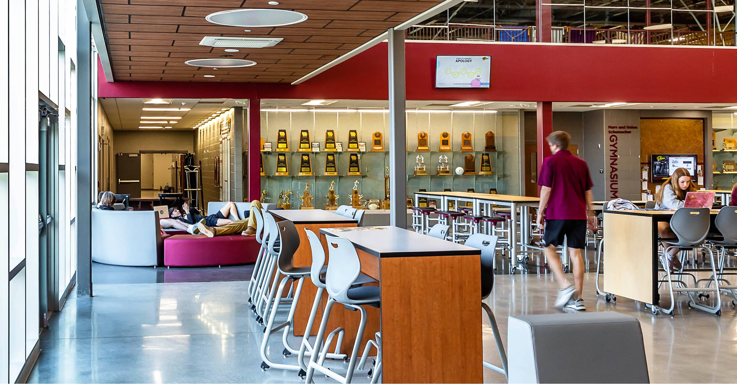 Modern, spacious lounge with wooden tables, white chairs, and a glass trophy case. Students relax and study, surrounded by bright natural light.