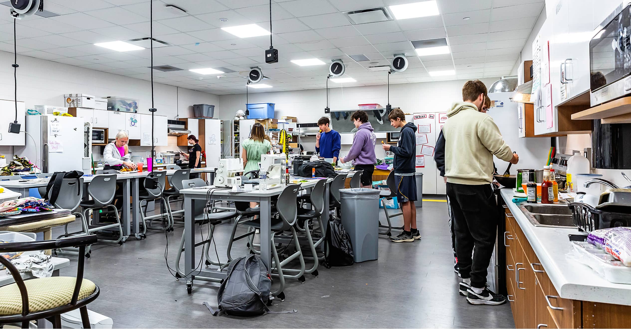 A busy classroom filled with students engaged in various activities. Some are working at high tables with computers and materials, while others are using the kitchen area. The room is well-lit, organized, and has a collaborative atmosphere.