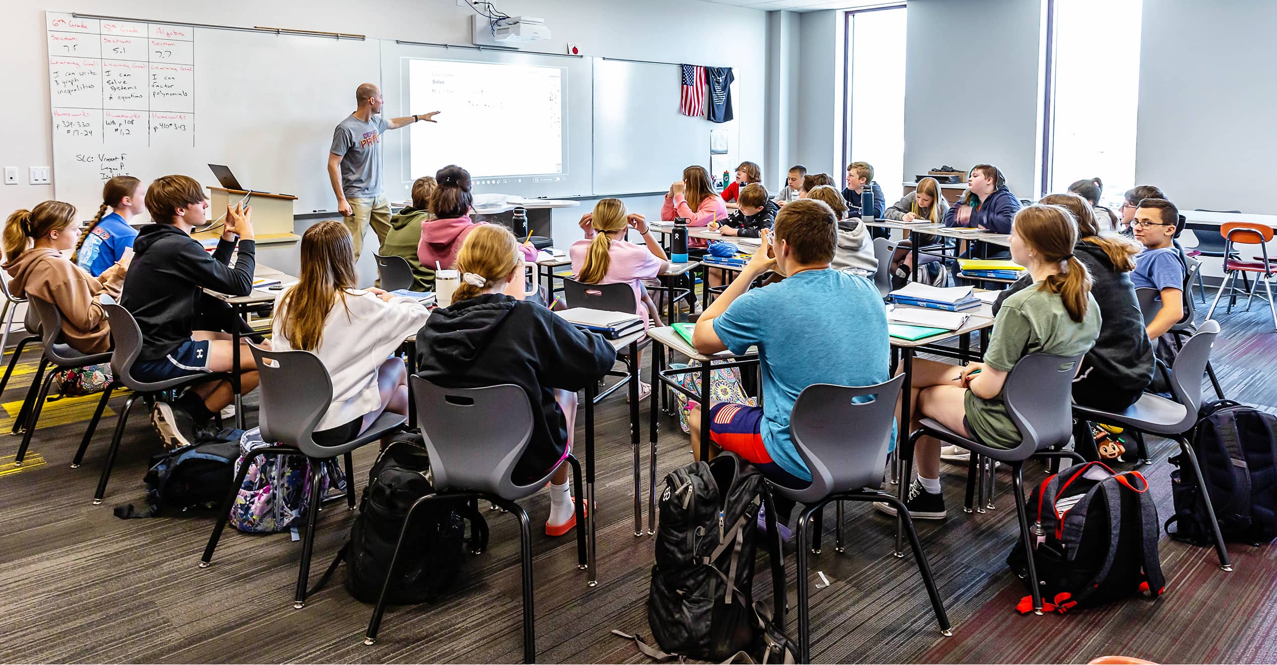 A classroom of students sits attentively around tables, facing a teacher pointing at a presentation on the whiteboard.
