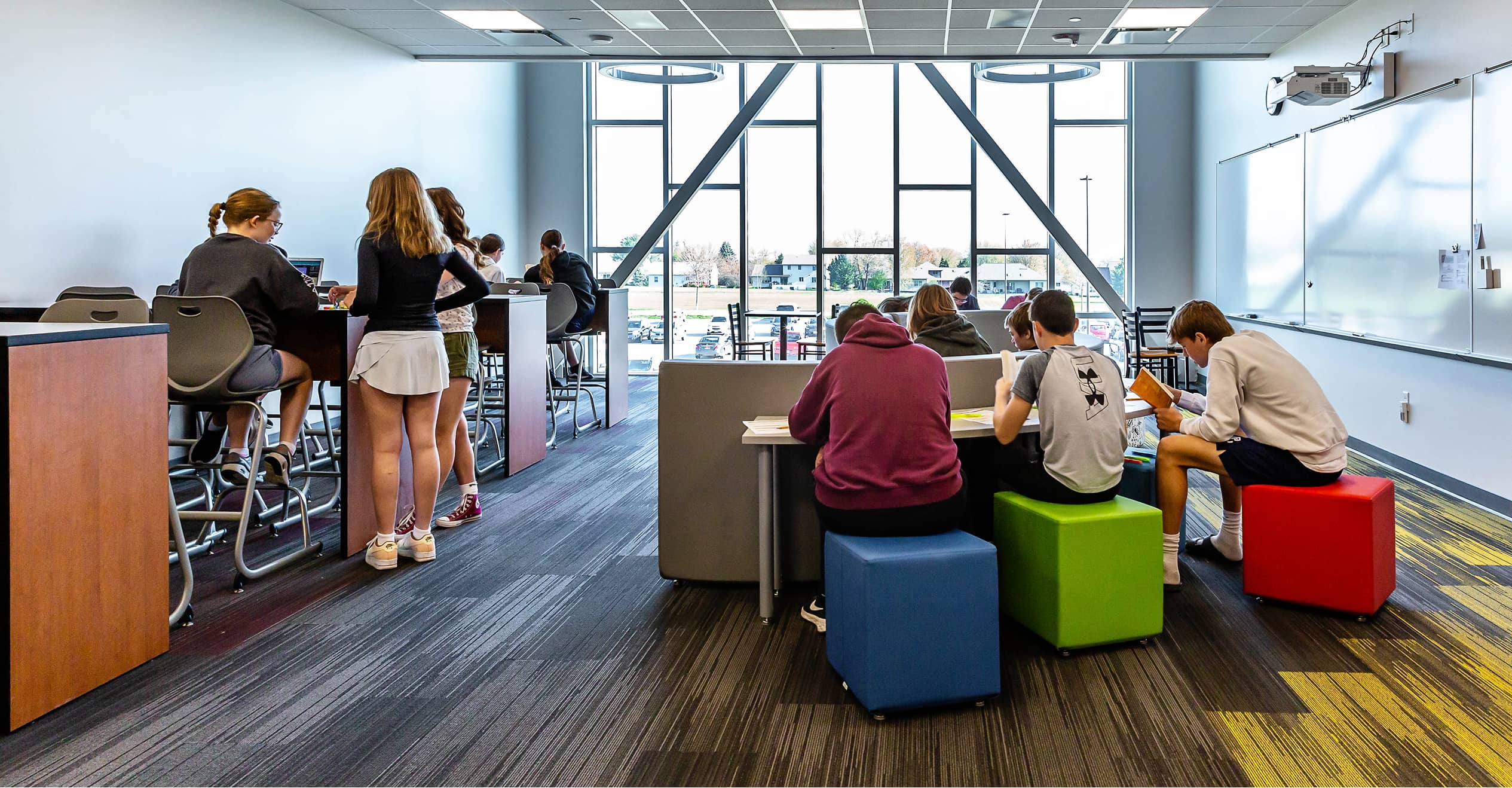 A modern classroom with students in groups, using colorful seating and tall desks. Large windows provide natural light