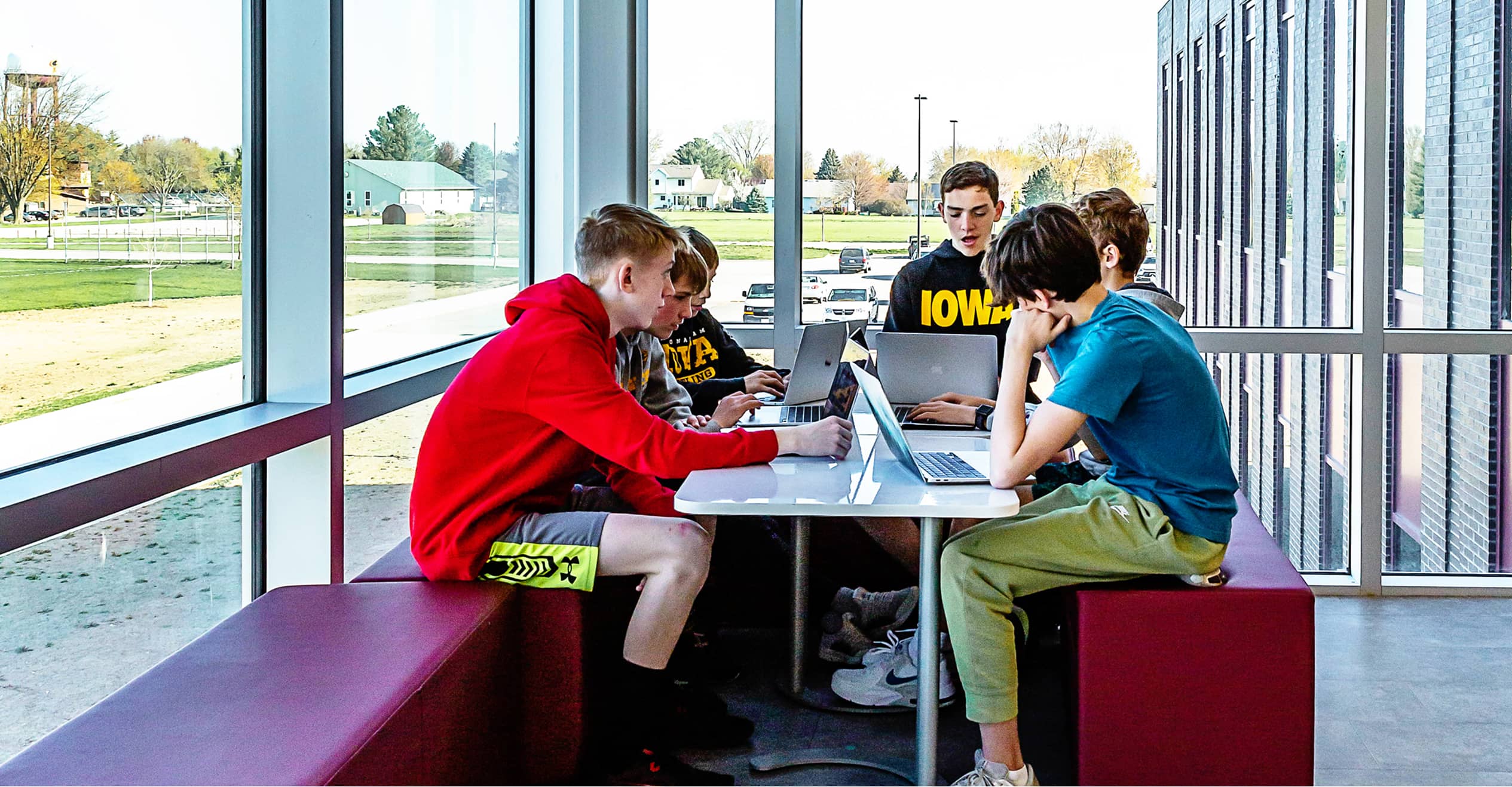 A group of five students sit at a table by large windows, focused on laptops. Their expressions are attentive and concentrated