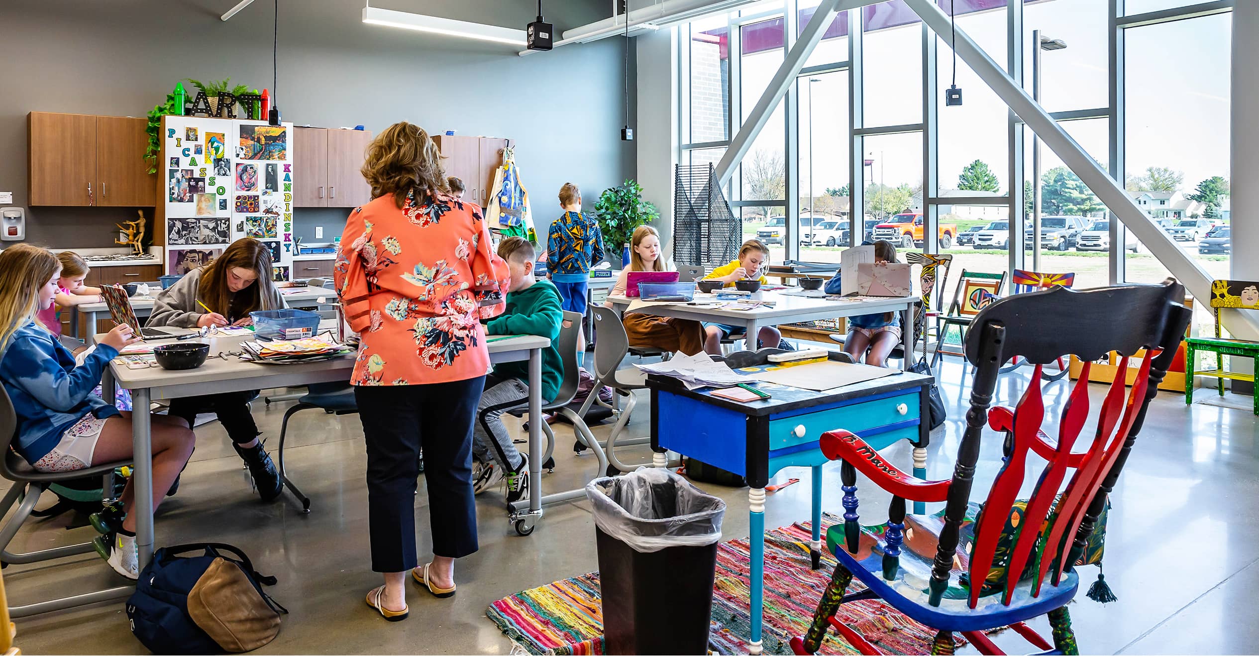 A classroom with students engaged in creative activities. A teacher in a floral blouse observes.