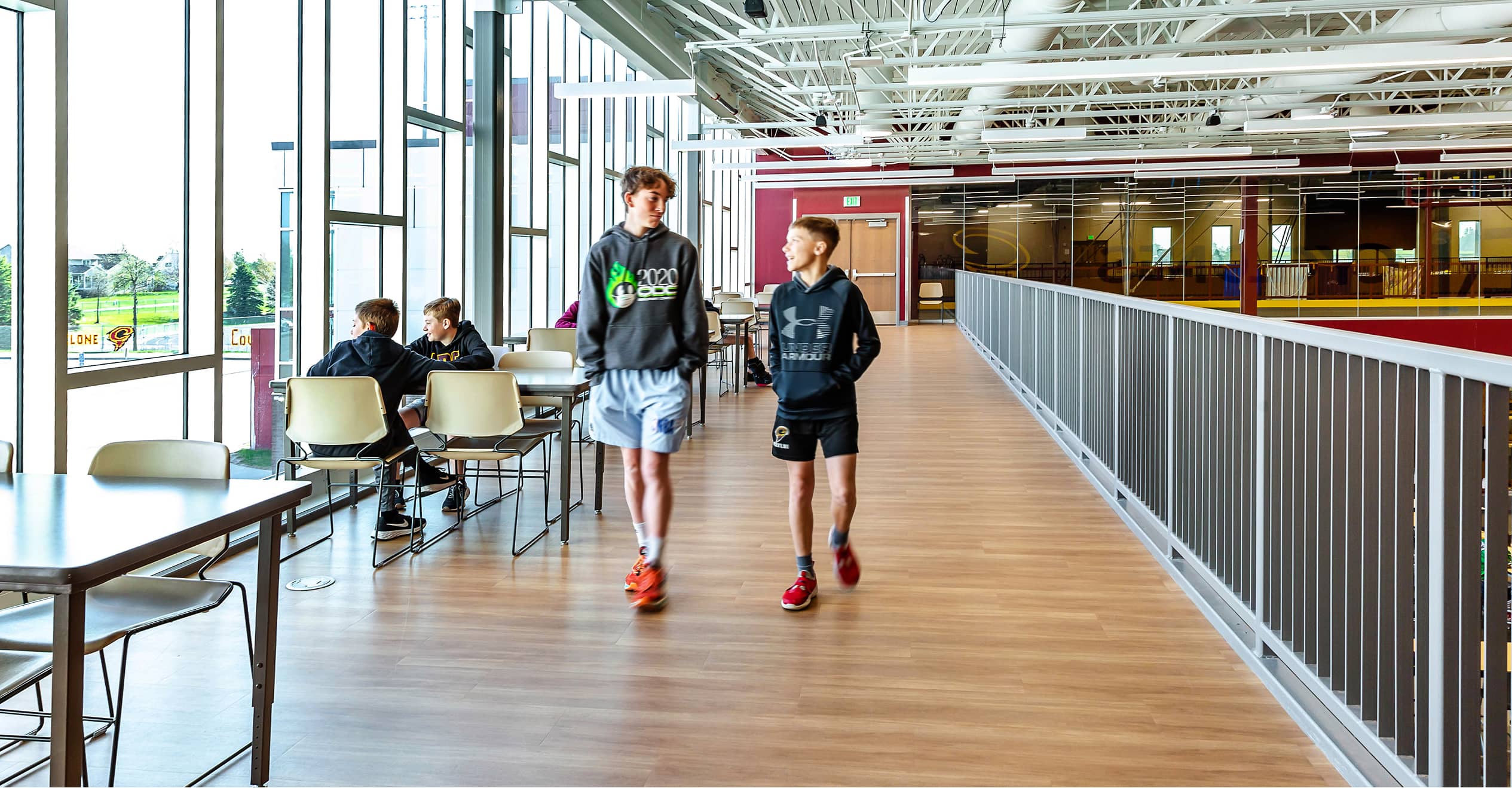 Two young people walk along a corridor with floor-to-ceiling windows, wearing casual sportswear. Others sit at tables, suggesting a relaxed, indoor setting.