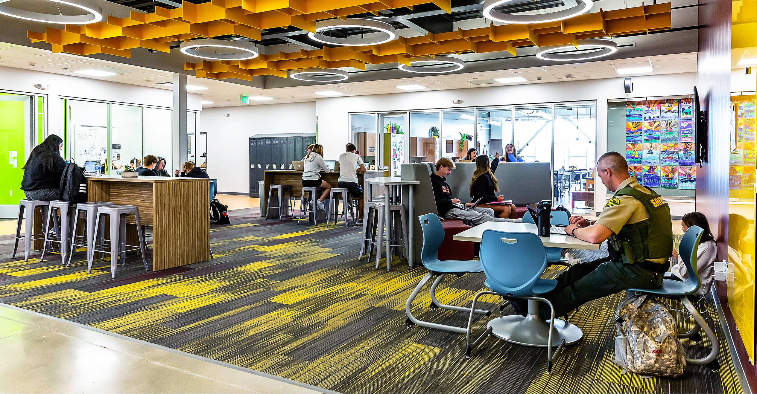 Students study in a modern library with colorful carpet, grey and blue furniture, and ring lights on the ceiling.