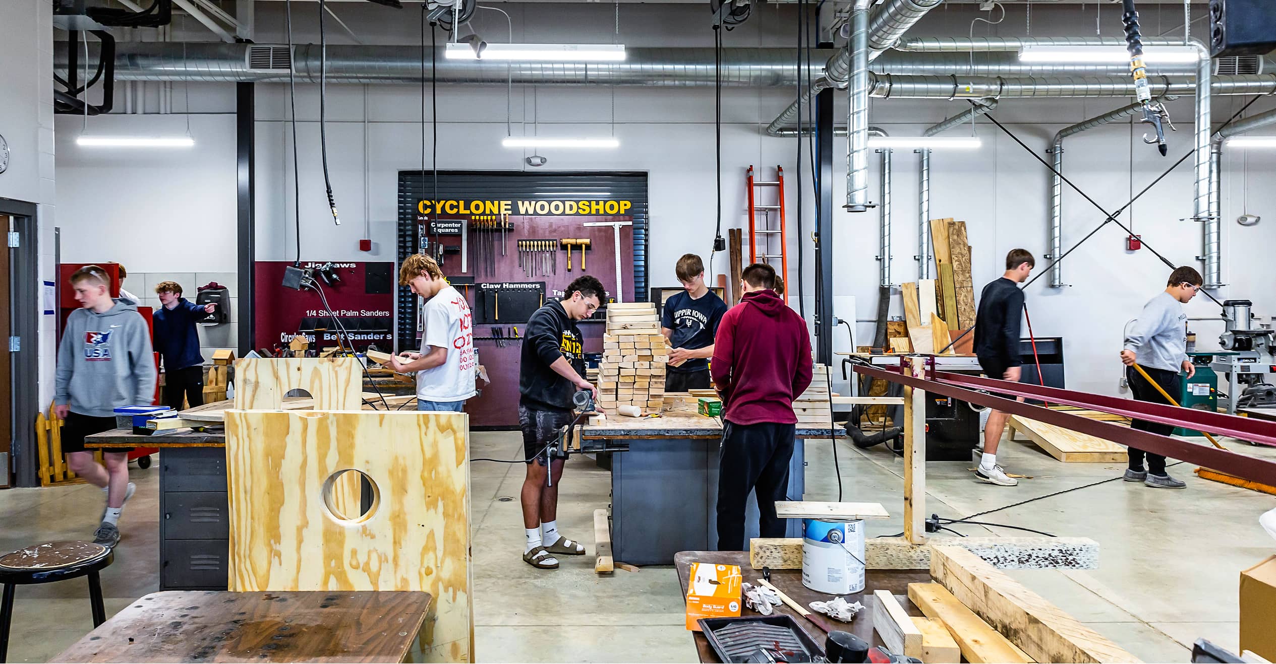 Students work in a busy school woodshop, crafting projects with various tools. The space is vibrant and active