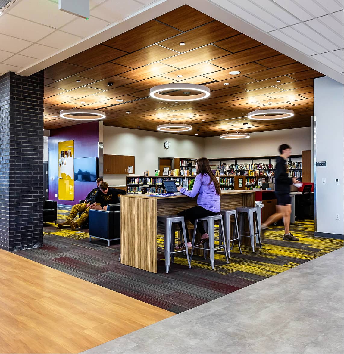 Modern library with wooden ceiling and circular lights. A person works on a laptop at a high table, while others sit nearby. Shelves of books line the walls.