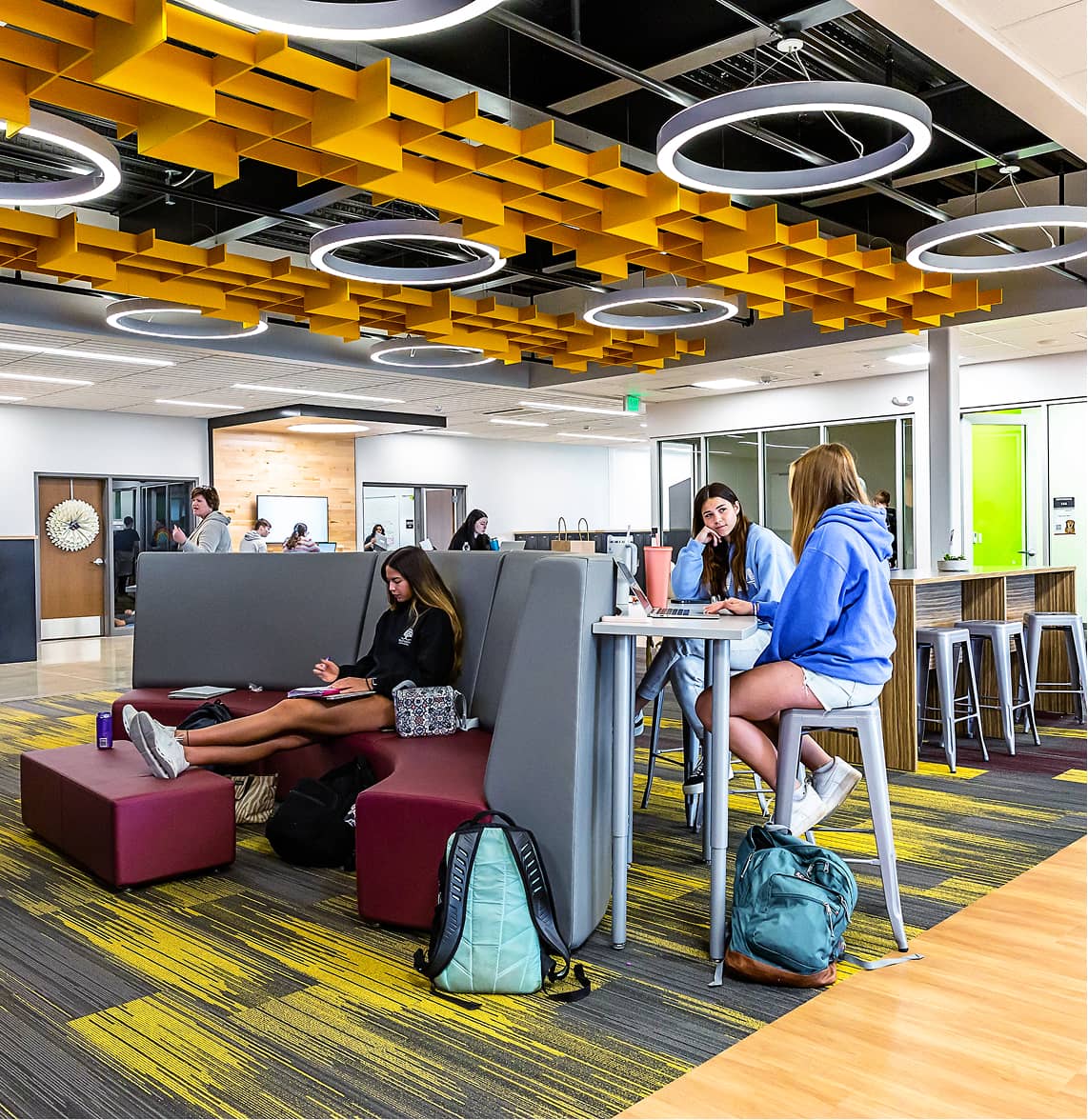 Modern, open study area with yellow ceiling accents. Students sit on gray and maroon furniture, engaging with laptops.