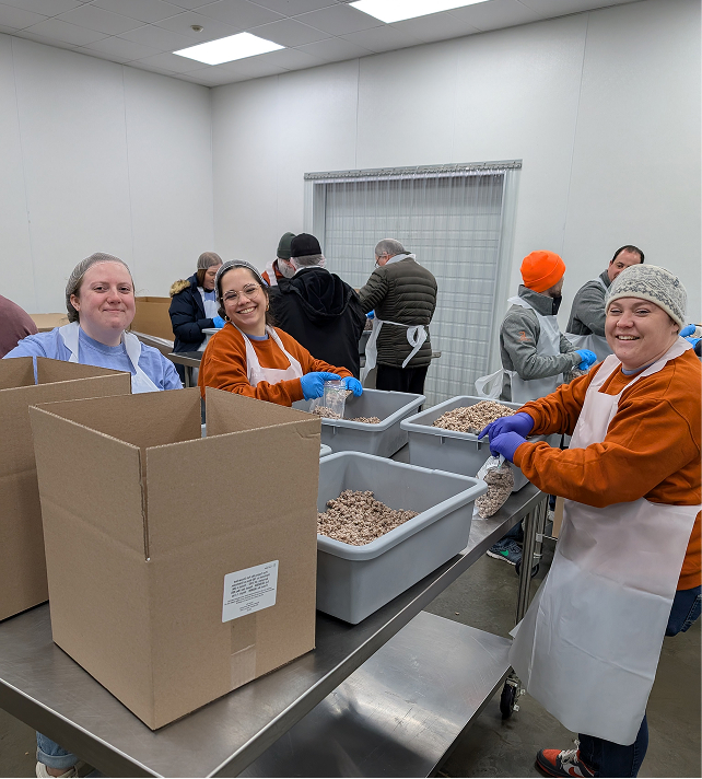 Smiling volunteers in a bright room pack dry food into containers. They wear aprons and gloves