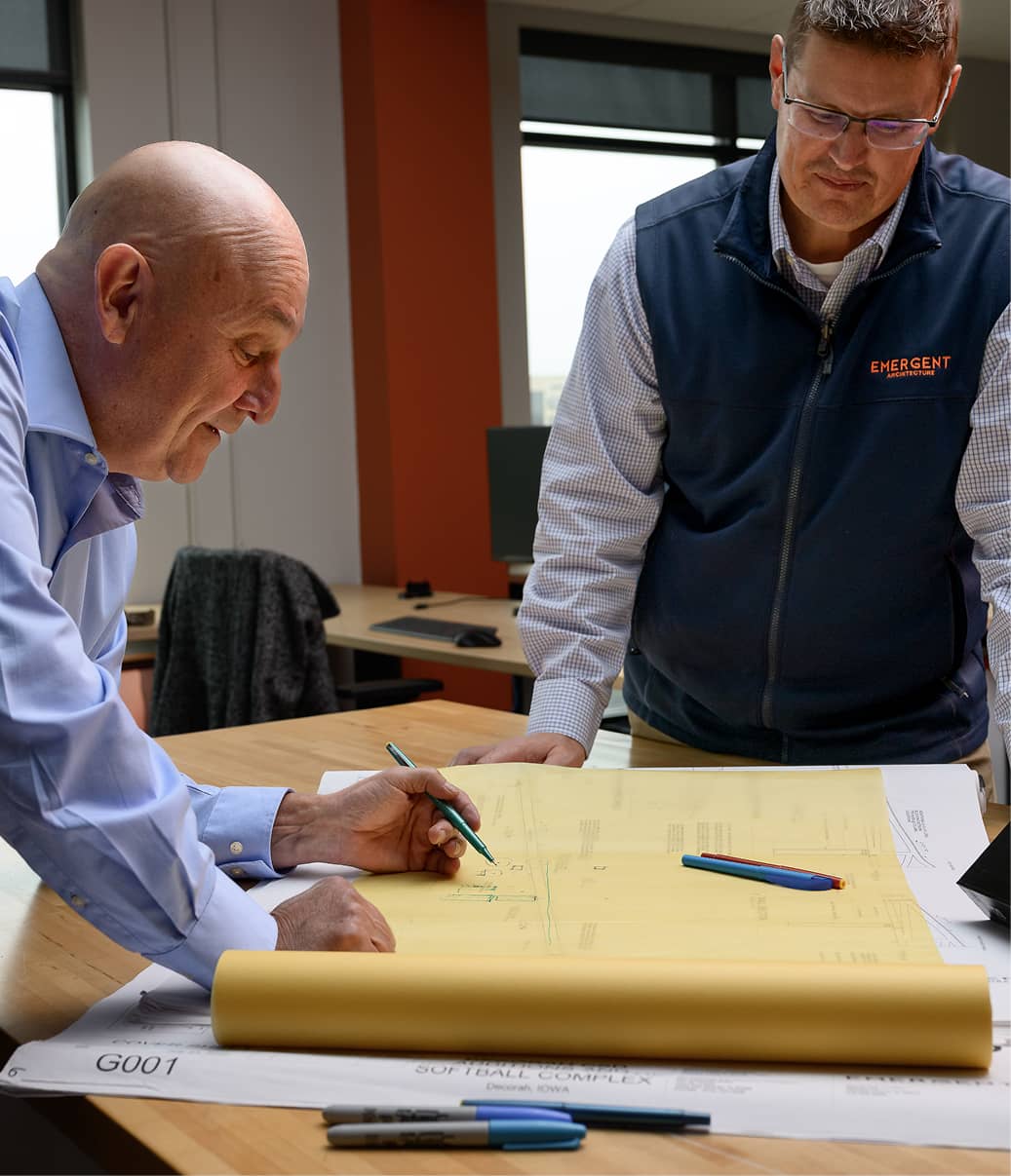 Two men are reviewing architectural plans on a desk in an office. One leans over, writing with a pen.