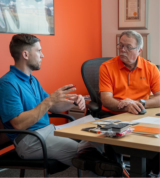 Two men engaged in conversation at a desk in an office. One wears a blue polo, gesturing as he speaks; the other listens, wearing an orange polo. Desk items include papers and a phone. Orange wall and framed certificates in background.