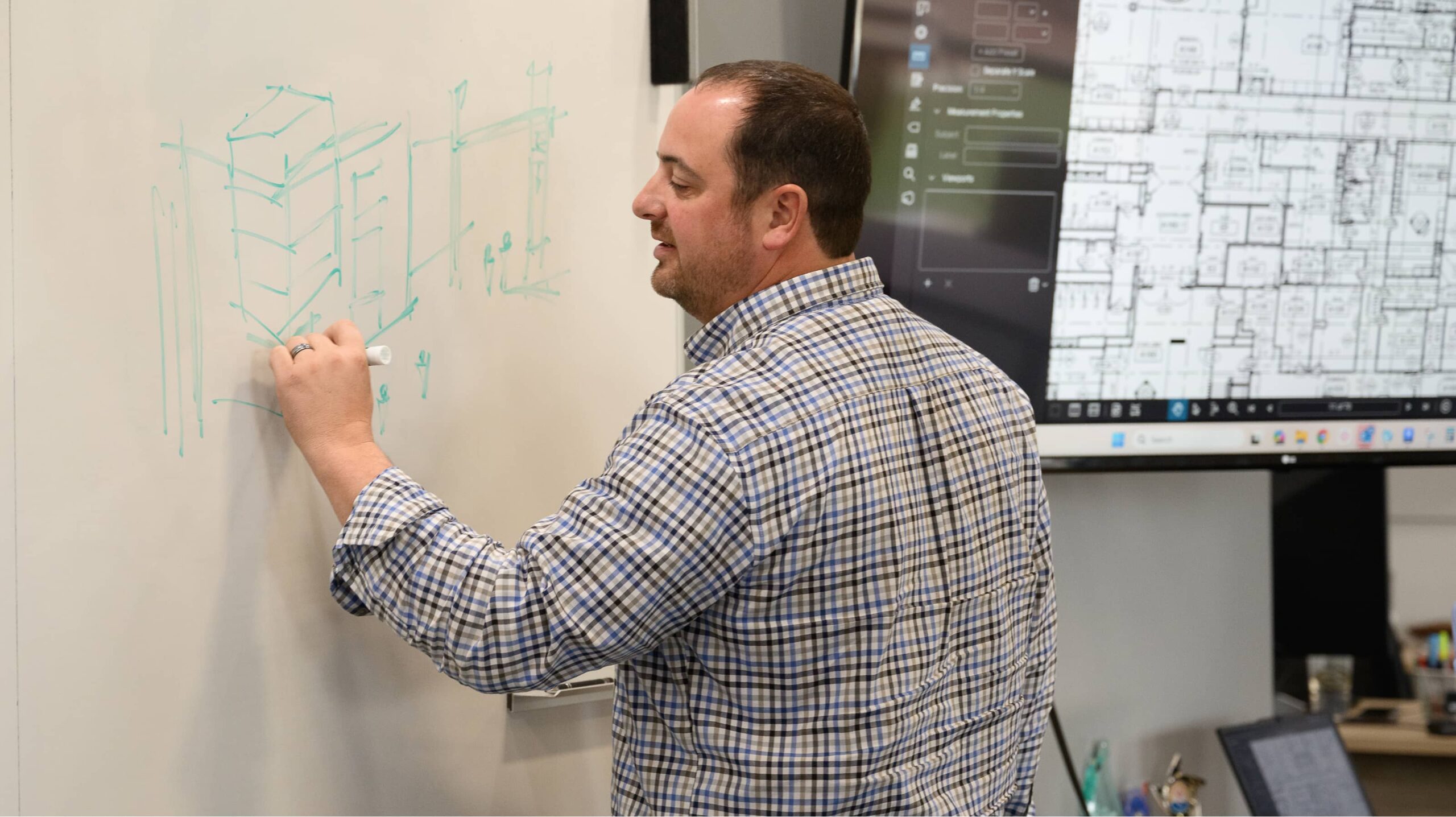 A man in a plaid shirt sketches architectural concepts on a whiteboard while building plans display on a monitor behind him at an architecture firm.