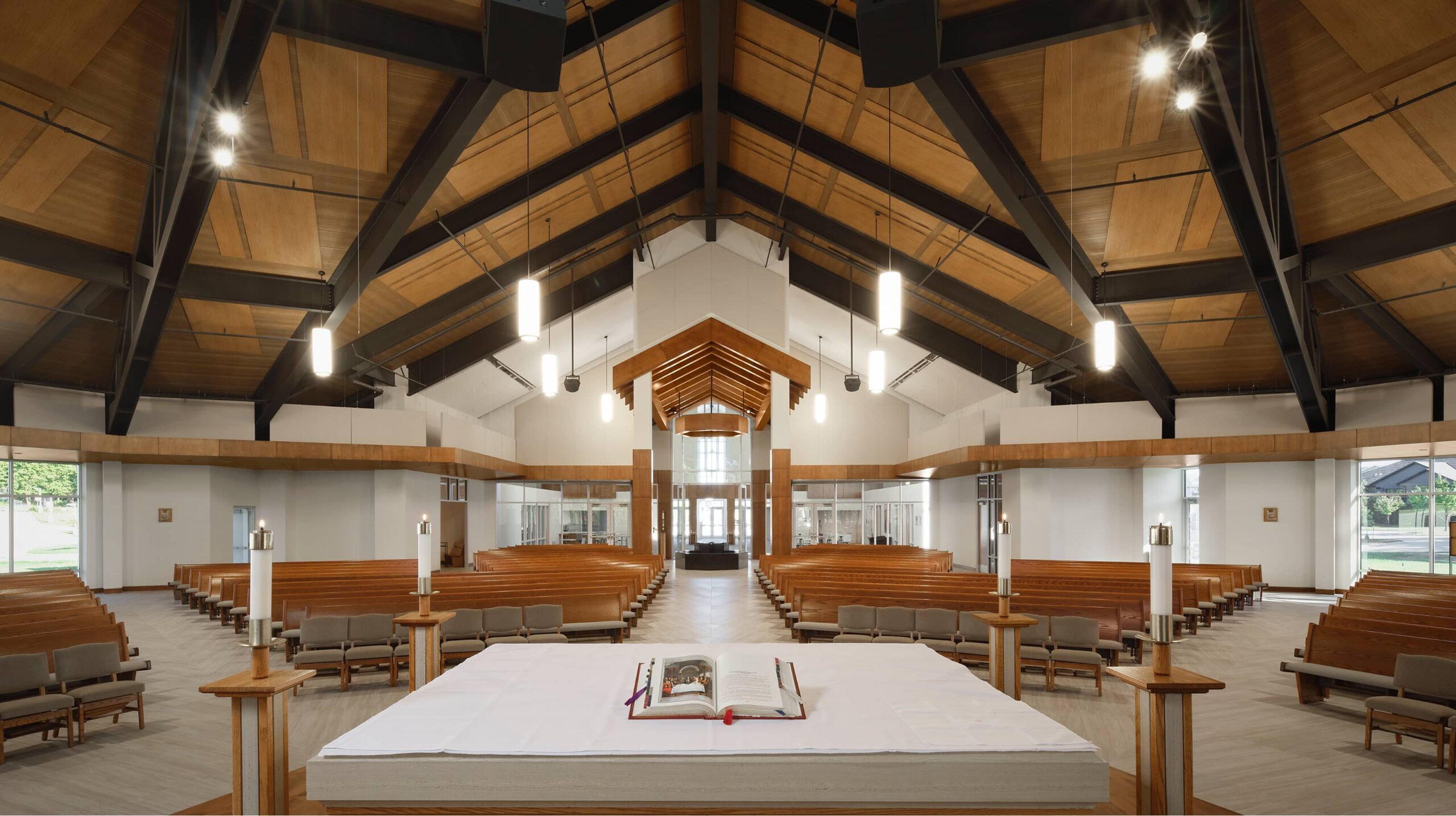 A modern church interior featuring a large altar with an open book, wooden pews arranged in a semicircle, and high, wooden-beamed ceilings with hanging lights.