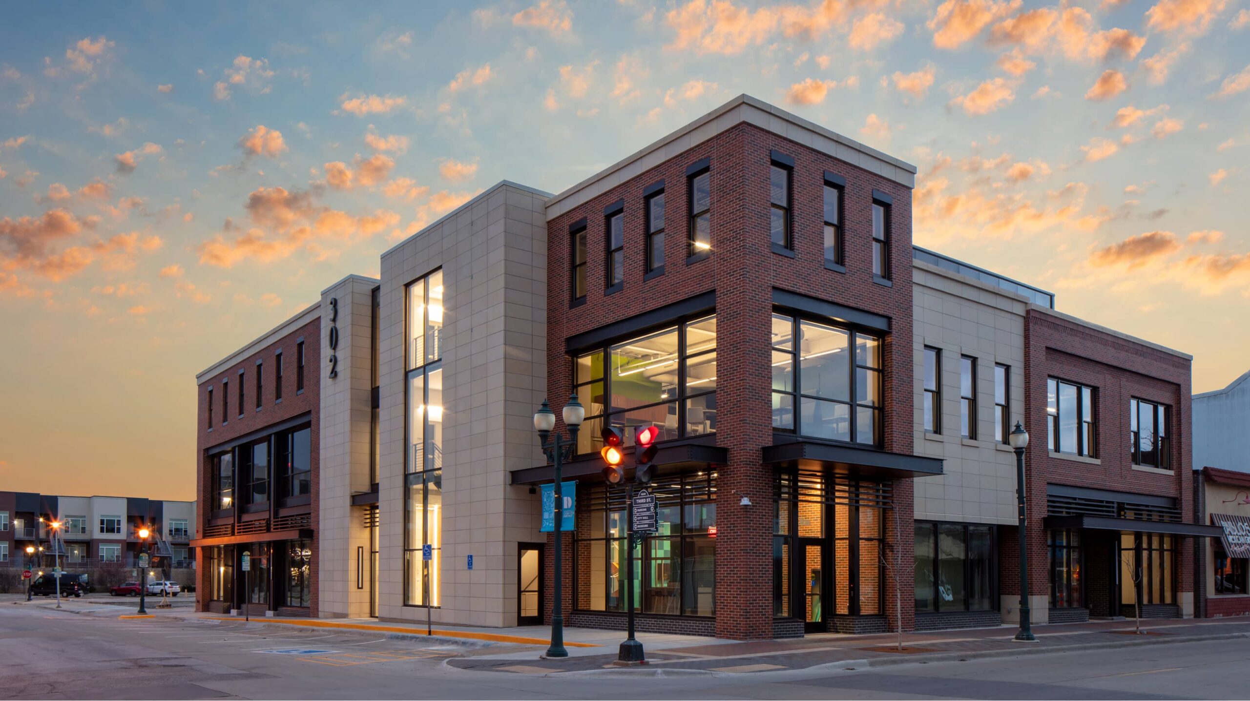 A modern two-story building designed by an architecture firm features brick and light-colored panels at a street corner during sunset under a partly cloudy sky.