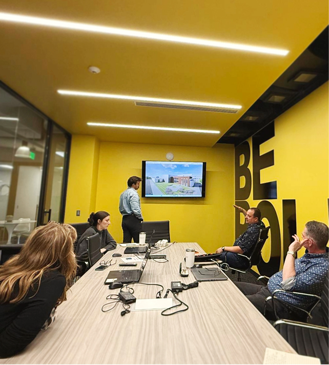 Meeting room with a yellow wall features "BE BOLD." Four people sit around a table while one presents on a wall screen