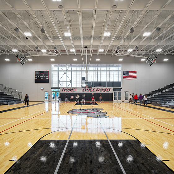 Bright, modern high school gymnasium featuring a wood basketball court, exposed ceiling structure, bleachers, and Home of the Bulldogs branding.