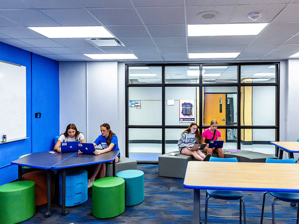 Classroom commons area with students seated at tables and flexible green stools.