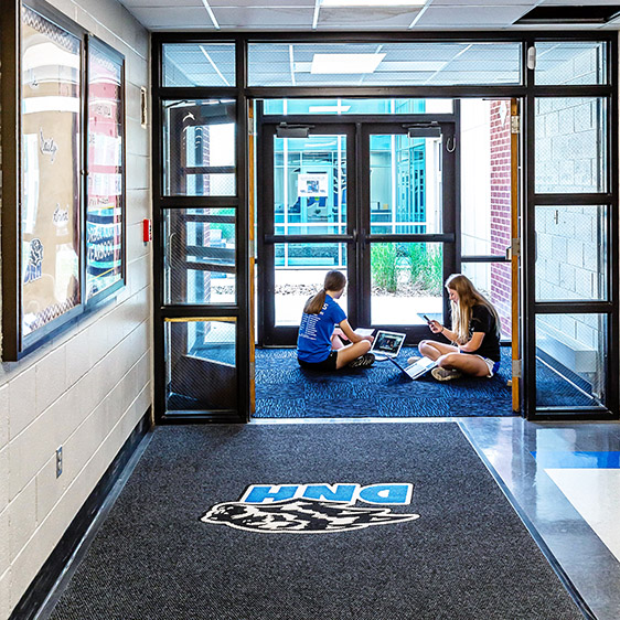 Students sitting and working in a bright school entry corridor with glass doors, natural light, and Dike-New Hartford branding on the floor.