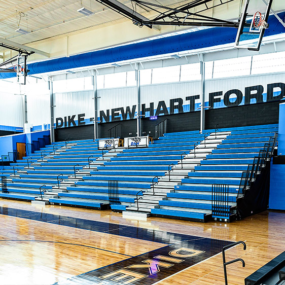 Interior view of the Dike-New Hartford High School gymnasium featuring blue bleachers, a polished wood court, and school branding along the back wall.