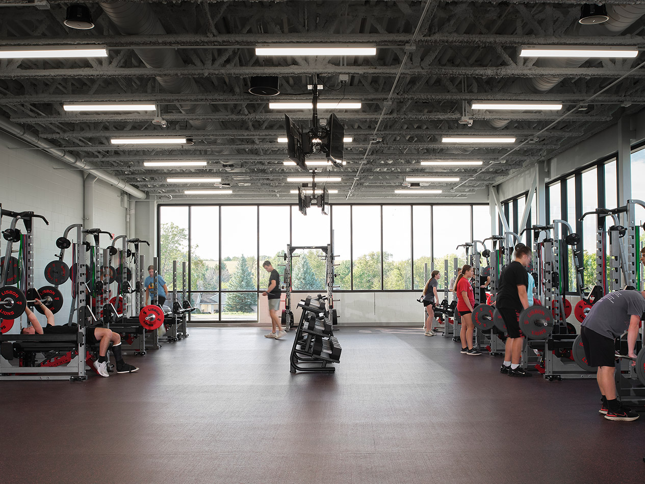 Student weight room with strength training equipment, free weights, and large windows providing natural light.