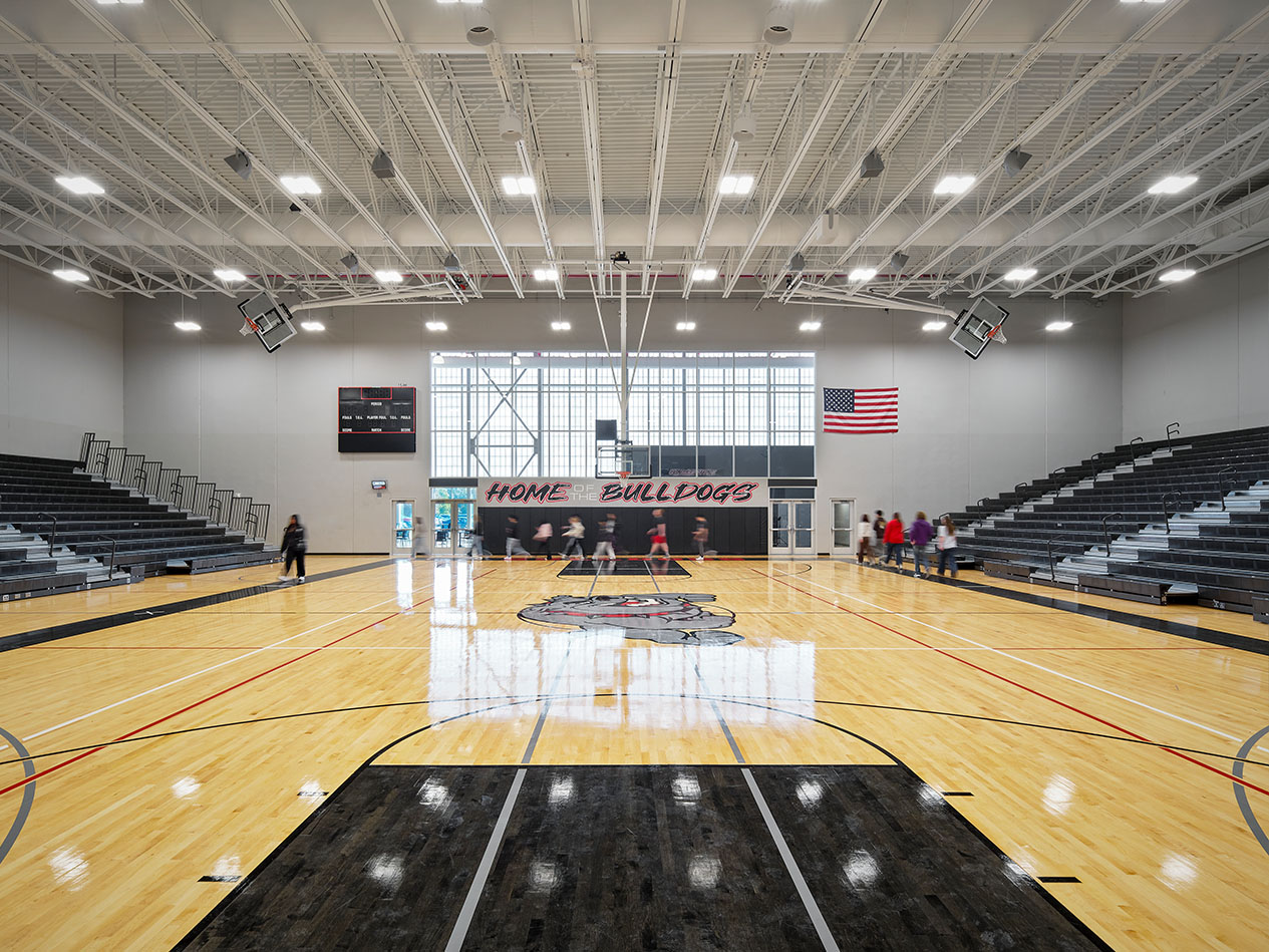 Modern school gym with center court logo, retractable bleachers, and bright ceiling lights.