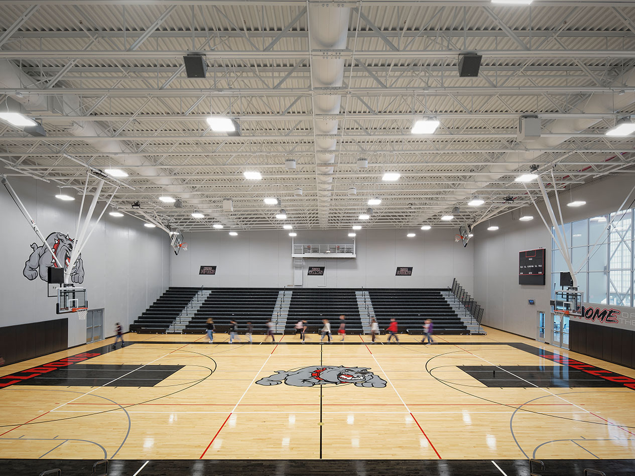 High school gymnasium with basketball court, bleachers, and overhead lighting.